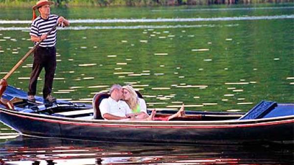 Couple kissing on a gondola with gondolier rowing on a green lake.